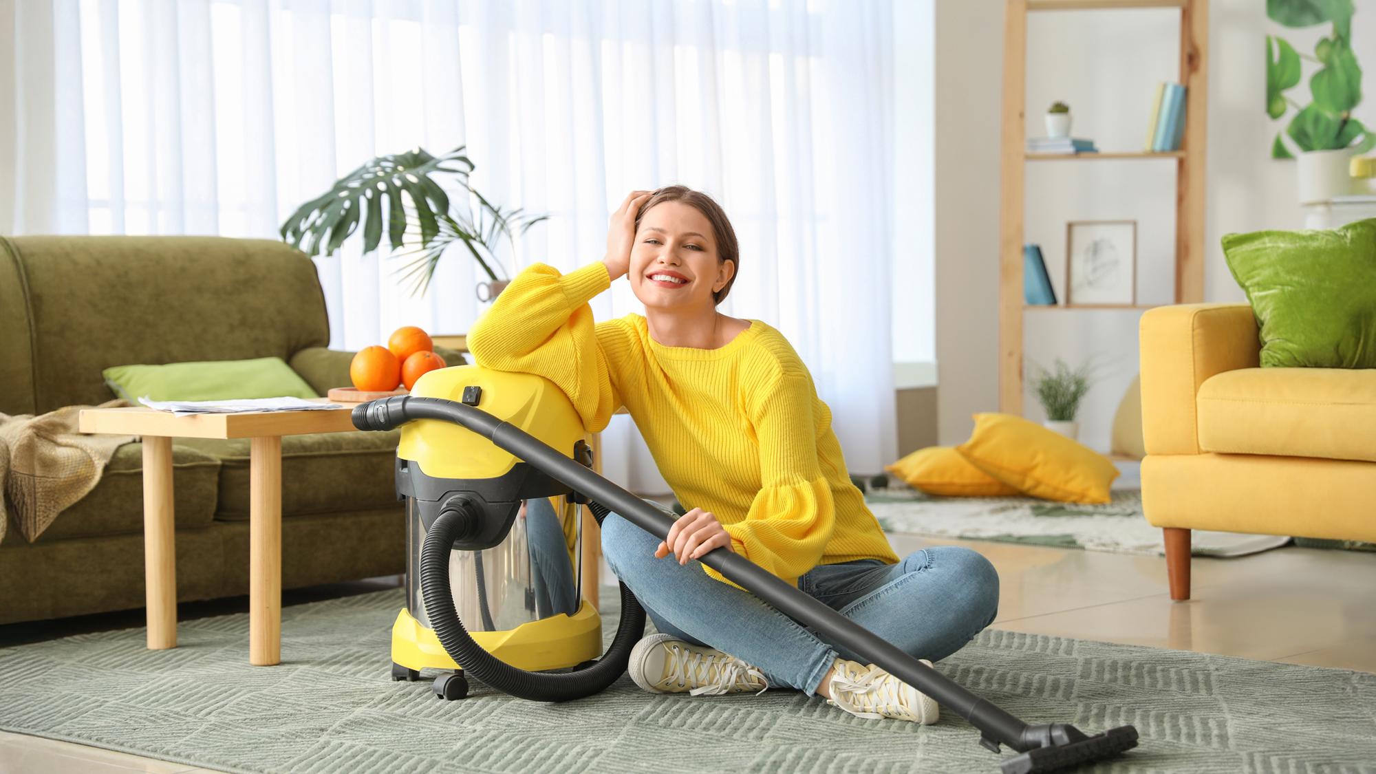 A young woman in a yellow sweater sitting on the floor next to a yellow vacuum cleaner, smiling cheerfully while leaning against it. The background features a bright room with plants and comfortable furniture.